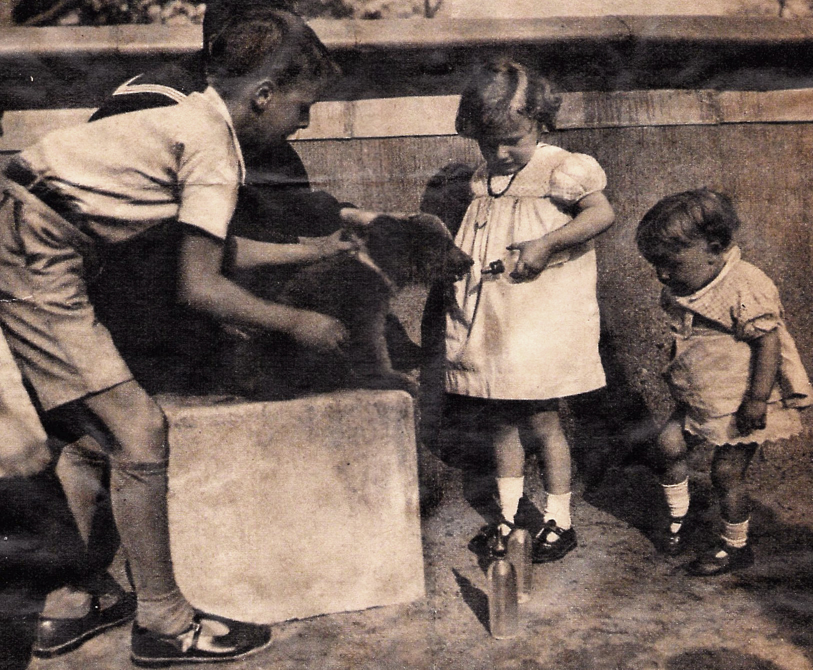 A bear cub sits on a stone; two children hold it by a neck band while two other children bottle feed it.