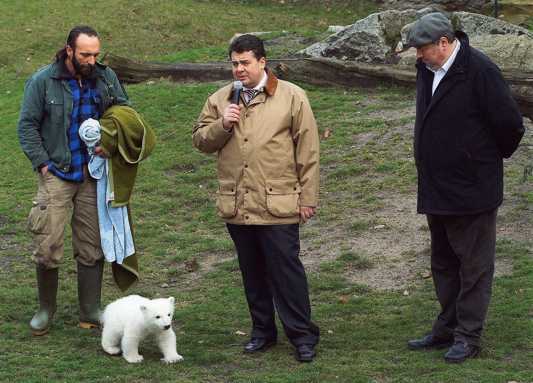 A bearded man on the left wearing the uniform and gumboots of a zookeeper stands on a lawn with a polar bear at his feet. At the centre, wearing a beige jacket, is a man with a microphone, to the right of him a man in dark clothing. The two people standing at the outside are turned towards the polar bear; the speaker is looking ahead.