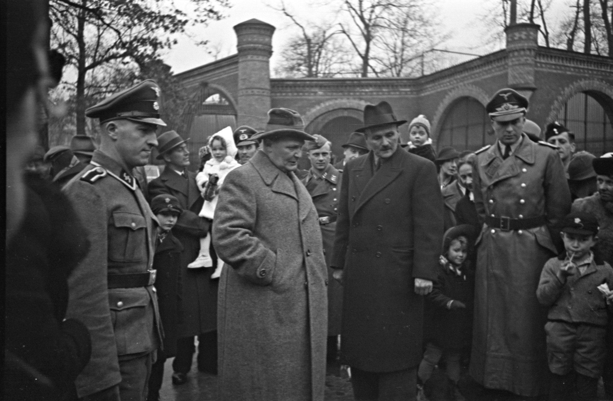 Black and white photograph: Two men in coats and hats surrounded by families with children and people in Nazi uniforms. Brick wall with window arches in the background.