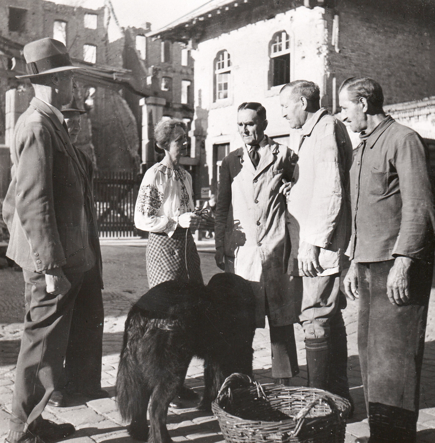 Black and white photograph: Four men, a woman, and a dog standing next to a woven basket, in front of partially destroyed buildings.