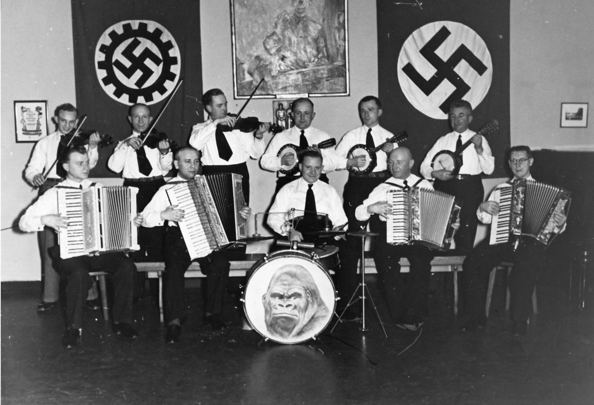 Black and white photograph: eleven people with string and percussion instruments and accordions. In the background are two large flags with swastikas and a painting. The bass drum displays a picture of a gorilla’s face.