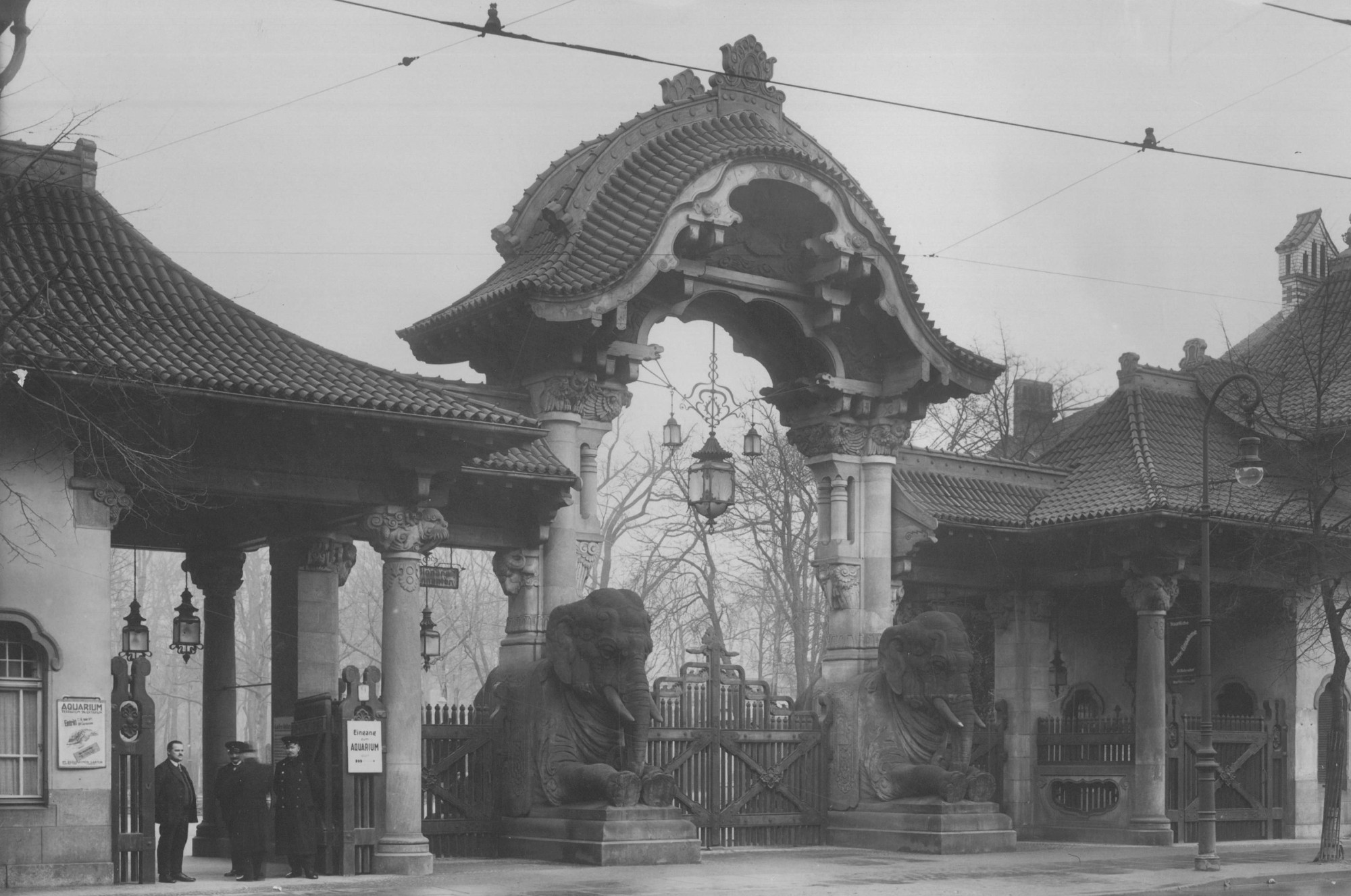 Black and white photograph: Gateway to Berlin Zoo supported by two elephant sculptures. Three men are standing in front of the entrance, two of them wearing zoo staff uniforms.