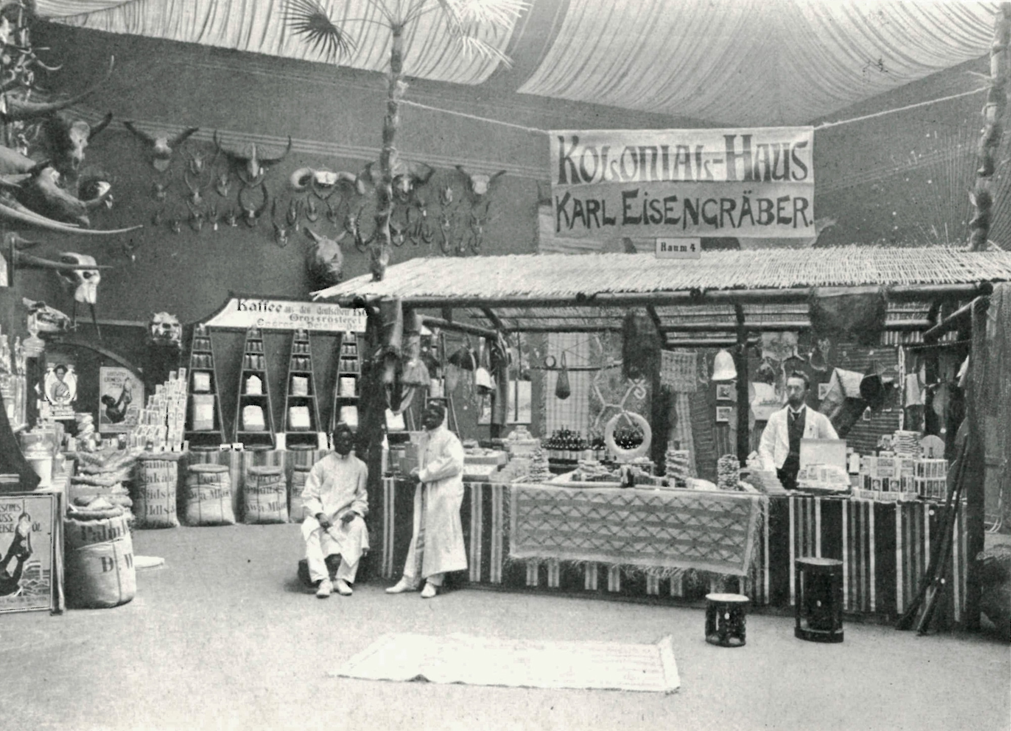 Black and white photograph of an exhibition room. Above a stand with various products like fabrics, crafted objects, and coffee, among other things, hangs a banner, advertising the “Kolonial-Haus Karl Eisengräber”. One white man is standing at the right corner behind the stand, two black men stand on the left side of the stand.
On the left wall are hanged animal skulls with horns of various sizes, below which are displayed several commercial products, such as coffee beans.