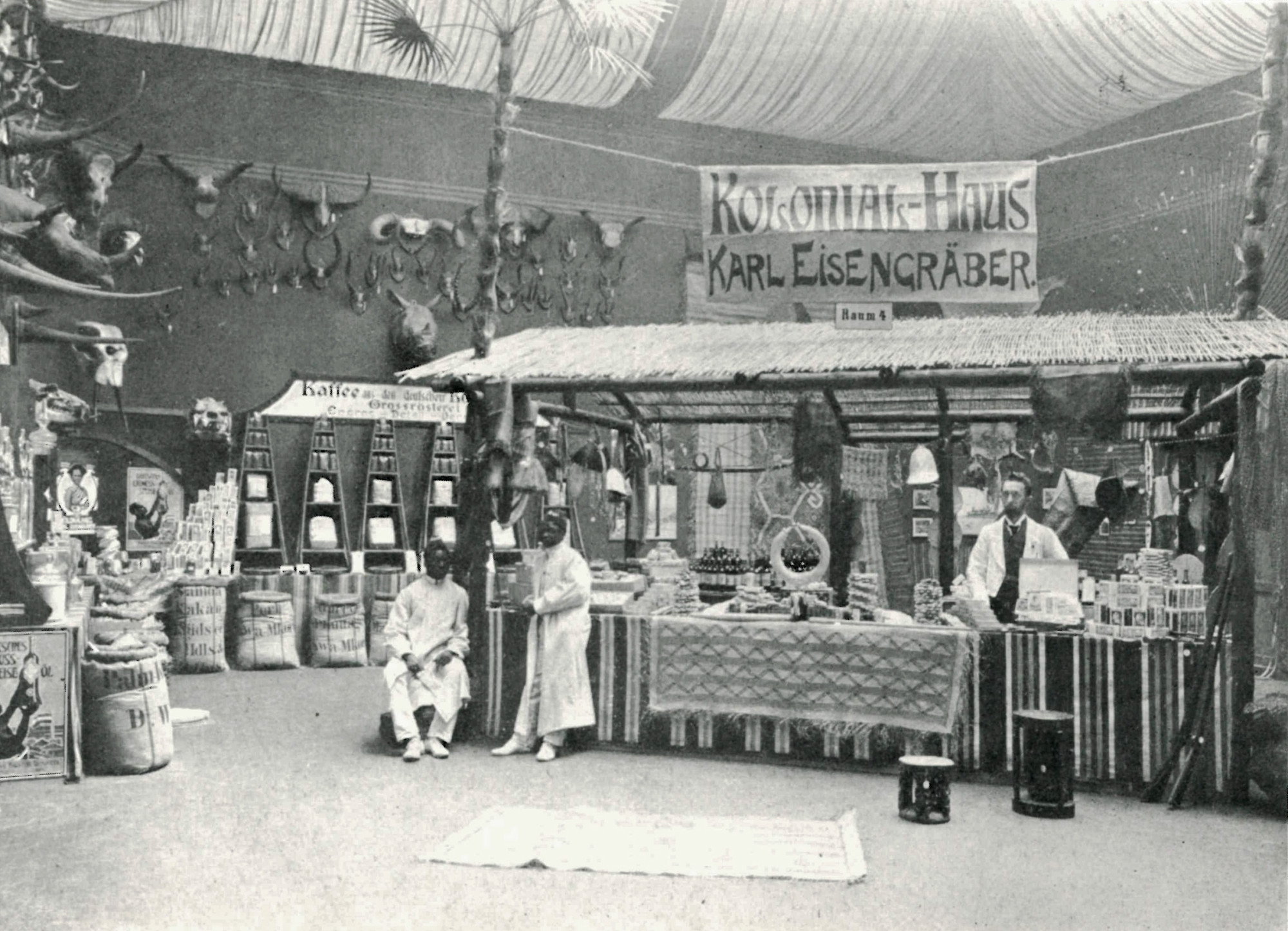 Schwarz-Weiß-Foto eines Ausstellungsraums. Oberhalb eines Verkaufsstands mit diversen Produkten wie Stoffen, handgemachten Gegenstände und Kaffee, hängt ein Banner mit der Aufschrift „Kolonial-Haus Karl Eisengräber“. Ein „Weißer“ Mann steht rechts im Bild hinter dem Verkaufsstand, am anderen Ende des Verkaufsstands befinden sich zwei „Schwarze“ Männer. An der Wand sind weitere Objekte zu sehen, so z.B. Kaffeeverpackungen, darüber hängen zahlreiche, unterschiedlich große, gehörnte Tierschädel.