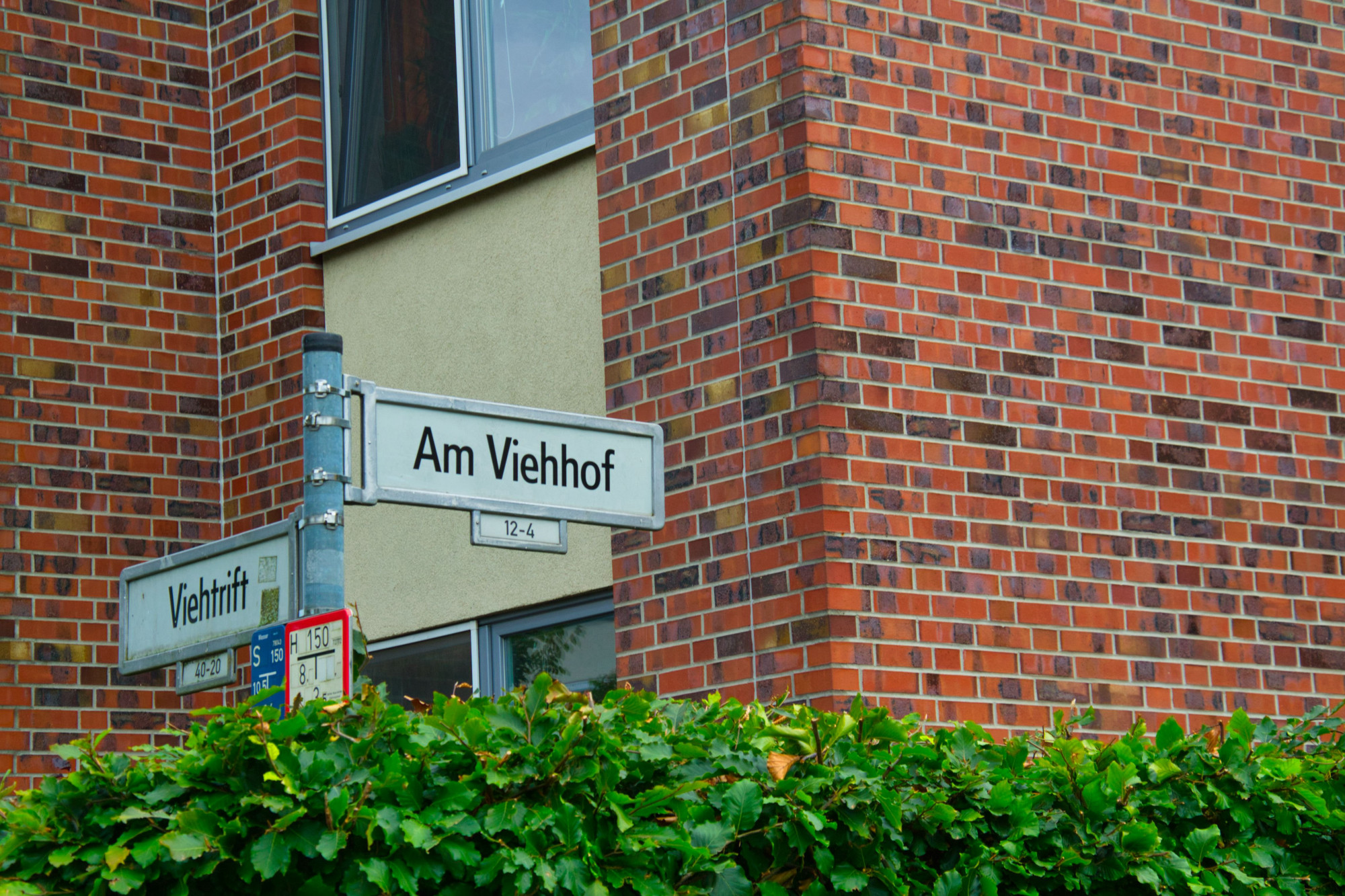 Fotografie einer Straßenecke. Auf einem Straßenschild sind zwei Straßennamen zu lesen: „Am Viehof“ und „Viehtrift“. Im Vordergrund ist eine grüne Hecke und im Hintergrund ein Backsteingebäude zu sehen.